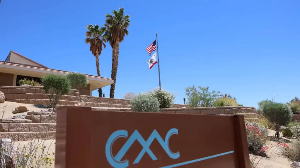 Copper Mountain College sign, palm trees, flagpole surrounded by plants and shrubs leading up to college entrance