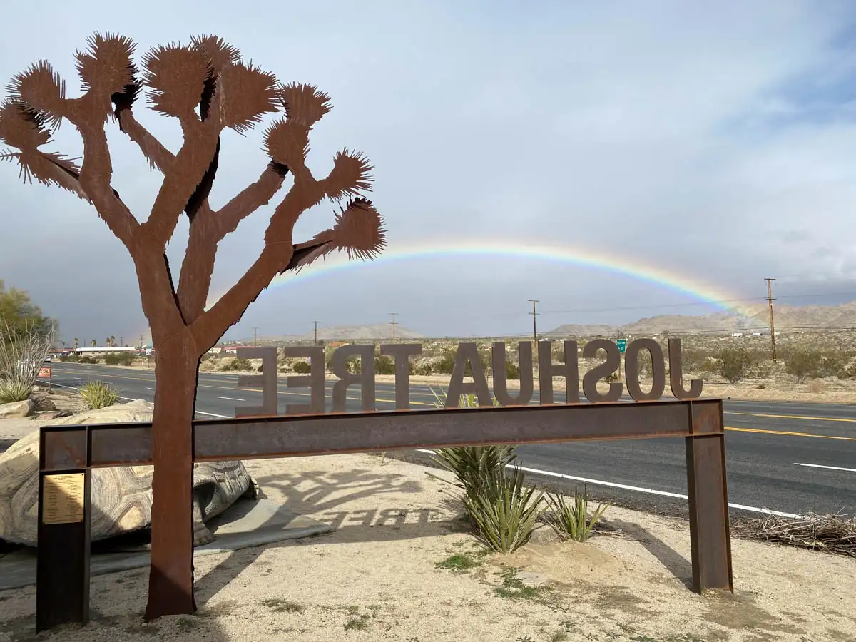 view of a full rainbow in front of the metal Joshua Tree sign on the freeway